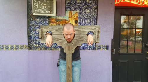 A person with a shaved head is playfully posing in a wooden stocks outside a colorful building.