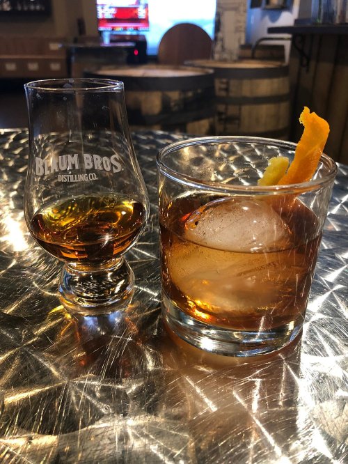 A glass of whiskey with an orange twist next to a smaller glass of whiskey on a reflective surface.