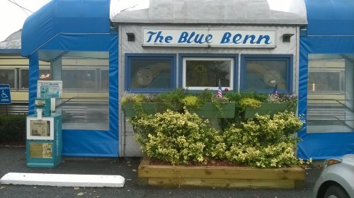 A blue and white diner with flower boxes and a menu stand outside, featuring the name "The Blue Benn."