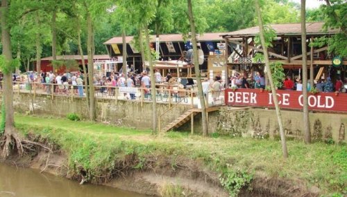 A lively outdoor bar scene by a river, with people enjoying drinks and dining under trees.