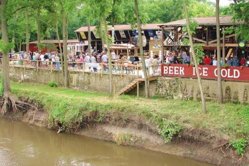 A lively riverside scene with people enjoying a bar and outdoor seating under trees.