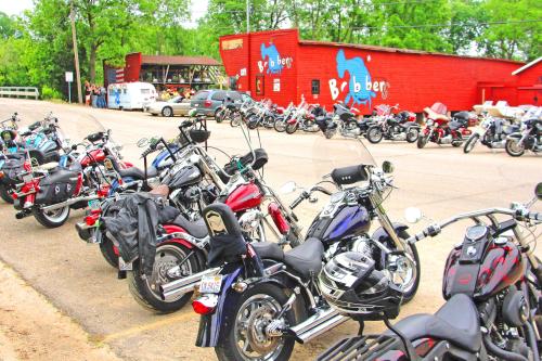 A row of parked motorcycles in front of a red building with a sign, surrounded by trees and other bikes.