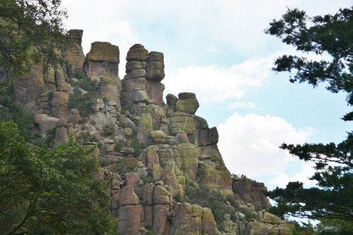Rocky cliff formation with tall, stacked boulders, surrounded by trees and a blue sky with scattered clouds.