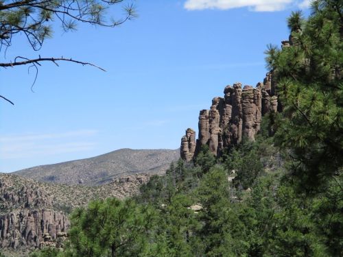 Rocky cliffs rise above a forested landscape under a clear blue sky, with distant mountains in the background.