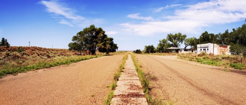 A deserted road stretches into the distance, flanked by sparse vegetation and an abandoned building under a clear blue sky.