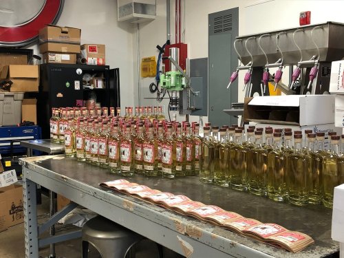 Bottles of liquor lined up on a table in a production facility, with labeling equipment and boxes in the background.