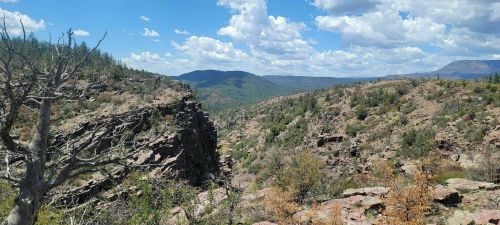 A scenic view of rocky hills and valleys under a blue sky with scattered clouds and greenery in the foreground.