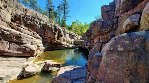 A serene rocky landscape with a calm pool of water surrounded by trees and cliffs under a clear blue sky.