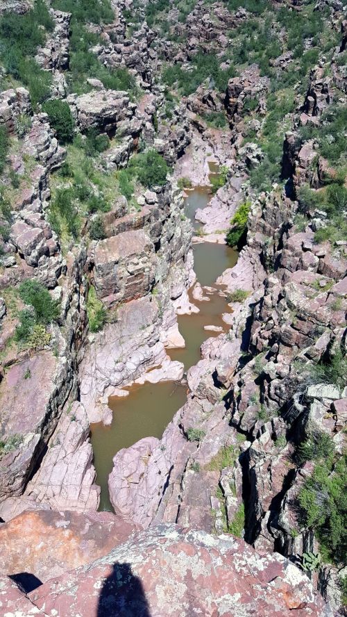 A narrow canyon with steep rock walls and a winding stream at the bottom, surrounded by greenery.