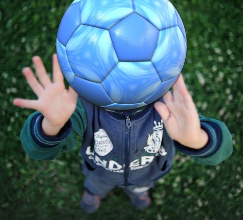 A child with a soccer ball for a head reaches out with both hands, standing on grass.