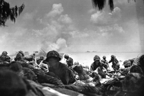Soldiers on a beach during a battle, with smoke rising in the background and a cloudy sky overhead.