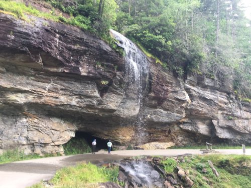 A scenic view of a waterfall cascading down a rocky cliff beside a walking path, with two people walking nearby.