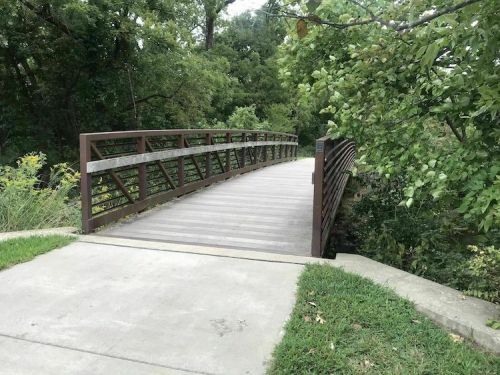 A wooden pedestrian bridge spans a small creek, surrounded by lush greenery and a paved pathway.
