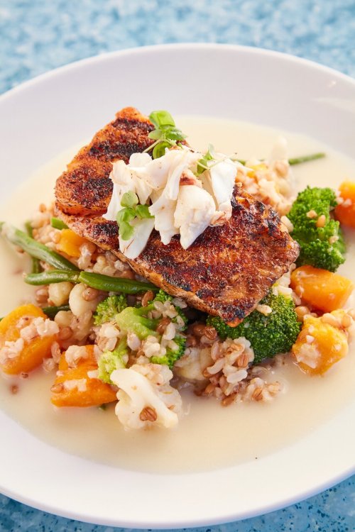 Grilled fish atop a bed of grains and mixed vegetables, garnished with crab and herbs in a creamy sauce.