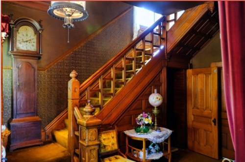 A vintage hallway featuring a wooden staircase, antique clock, and decorative table with flowers. Warm, inviting atmosphere.