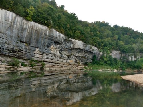 A serene river scene with a rocky cliff and lush greenery reflected in the calm water.