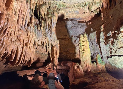Visitors explore a cave with impressive stalactites and stalagmites illuminated by soft lighting.