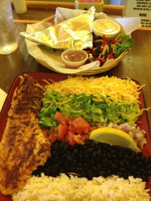 A colorful plate featuring grilled fish, rice, black beans, lettuce, tomatoes, cheese, and a lemon wedge, with a side salad.