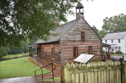 Historic wooden building with a bell tower, surrounded by greenery and a fence, on a rainy day.