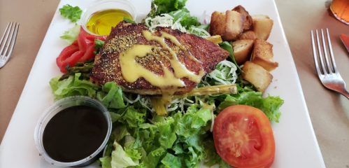 A plate of grilled fish on a bed of lettuce, topped with mustard sauce, served with tomatoes, potatoes, and dressings.