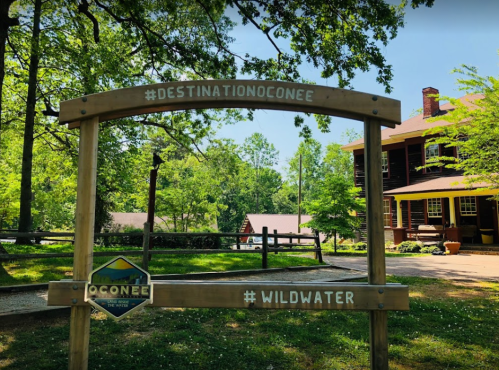 A wooden sign with hashtags #DESTINATIONOCONEE and #WILDWATER, surrounded by greenery and a rustic building in the background.