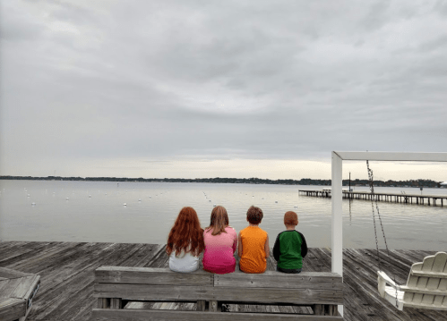 Four children sit on a dock, facing a calm lake under a cloudy sky, with boats visible in the distance.