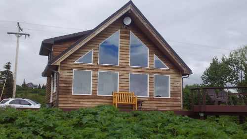 A wooden house with large windows and a deck, surrounded by greenery under a cloudy sky.