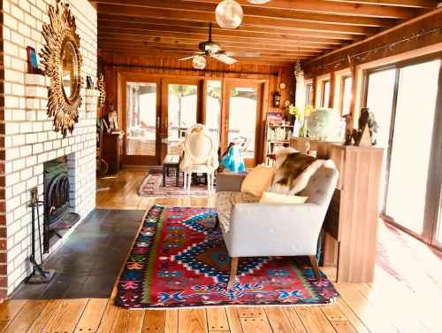 Cozy living room with wooden beams, a fireplace, colorful rug, and large windows letting in natural light.