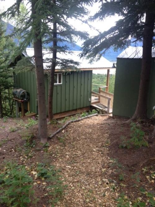 A path leads to a green cabin nestled among trees, with mountains visible in the background.