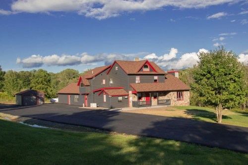 A large, modern house with red accents, surrounded by greenery and a clear blue sky. A paved driveway leads to the entrance.