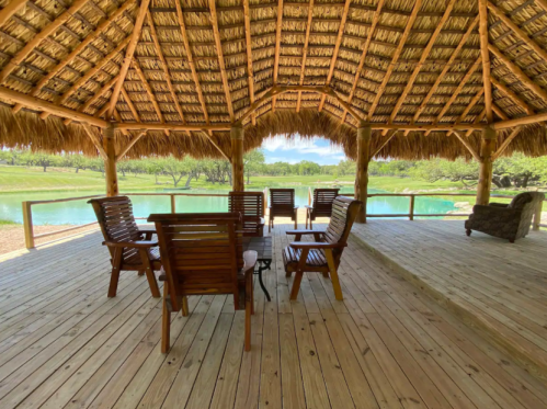 A spacious wooden deck with chairs overlooking a serene lake and lush greenery under a thatched roof.