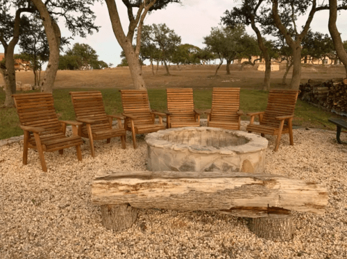 A circular stone fire pit surrounded by wooden chairs on a gravel area, with trees in the background.
