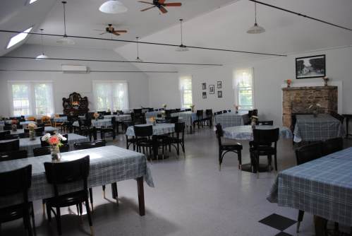 A spacious dining hall with tables set for meals, featuring checkered tablecloths and a stone fireplace.