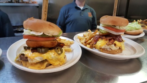 Two large burgers with toppings, served with fries on a plate, in a diner setting.