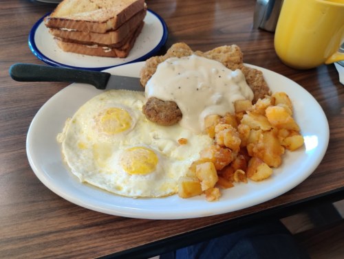 A plate of breakfast featuring two fried eggs, chicken fried steak with gravy, hash browns, and toast.