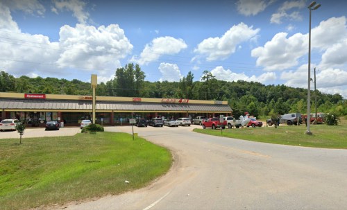 A strip mall with various storefronts, surrounded by green trees and a cloudy blue sky. Cars are parked in front.