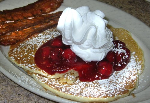 A plate of pancakes topped with cherry sauce and whipped cream, alongside crispy bacon strips.