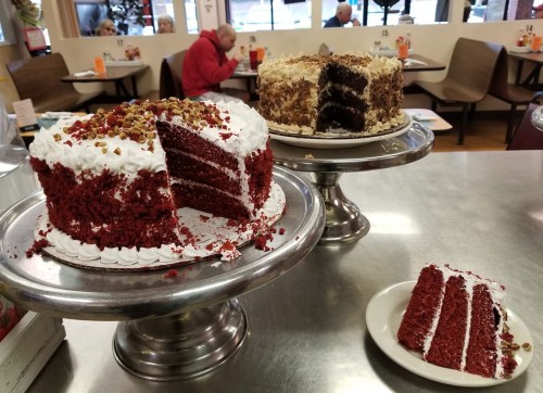 Three cakes on display: a red velvet cake with a slice missing, a layered chocolate cake, and a slice on a plate.