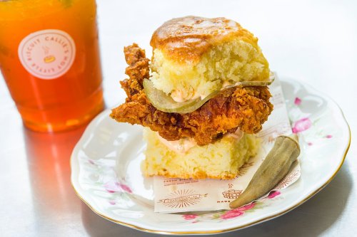 A biscuit sandwich with fried chicken, pickles, and sauce, served on a floral plate with a glass of iced tea.