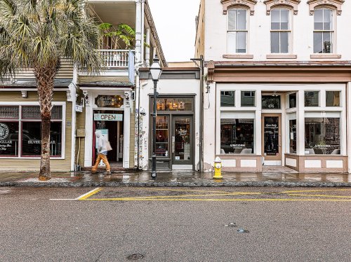 A street view featuring shops with open signs, palm trees, and a person walking by on a rainy day.