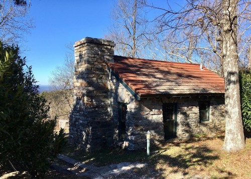 A small stone cottage with a red roof, surrounded by trees and a clear blue sky.