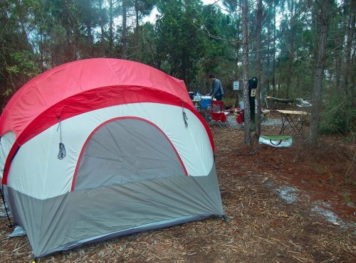 A red and gray tent set up in a wooded campsite with a person preparing food at a nearby table.