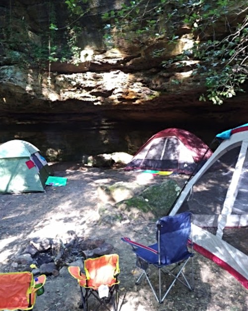 A campsite with colorful tents set up near a rocky overhang, surrounded by trees and a small fire pit.