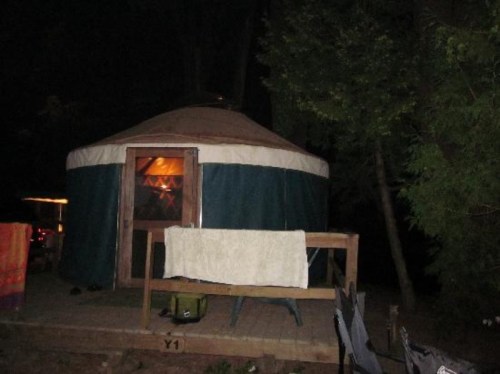 A yurt at night, with a wooden deck and a towel hanging on the railing, surrounded by trees.