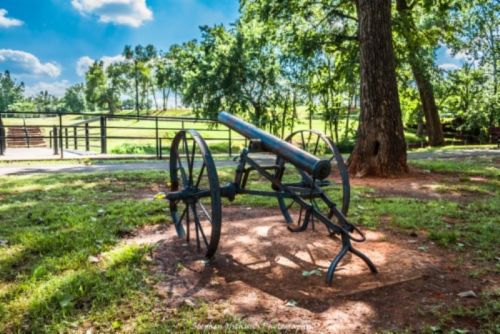 A historic cannon on a grassy area, surrounded by trees and under a blue sky with fluffy clouds.