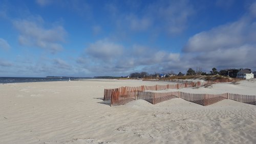 A sandy beach with wooden fencing, under a blue sky with clouds, and a distant shoreline.