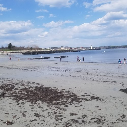 A sandy beach with people walking along the shore, under a blue sky with clouds and a distant lighthouse.