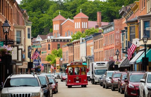 A charming street lined with historic buildings, parked cars, and a red trolley, with greenery and a church in the background.