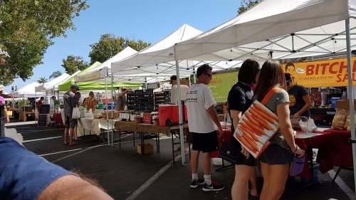 A bustling outdoor market with white tents, vendors, and shoppers browsing various goods and produce.