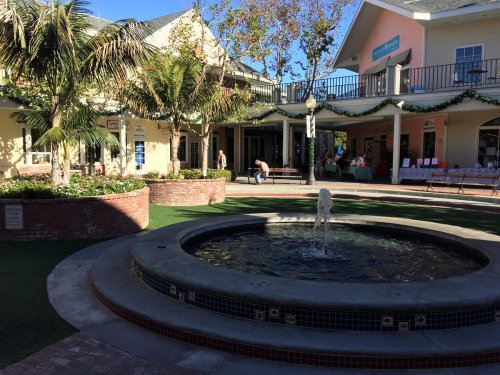 A sunny plaza with a fountain, surrounded by shops and greenery, featuring people enjoying the outdoor space.